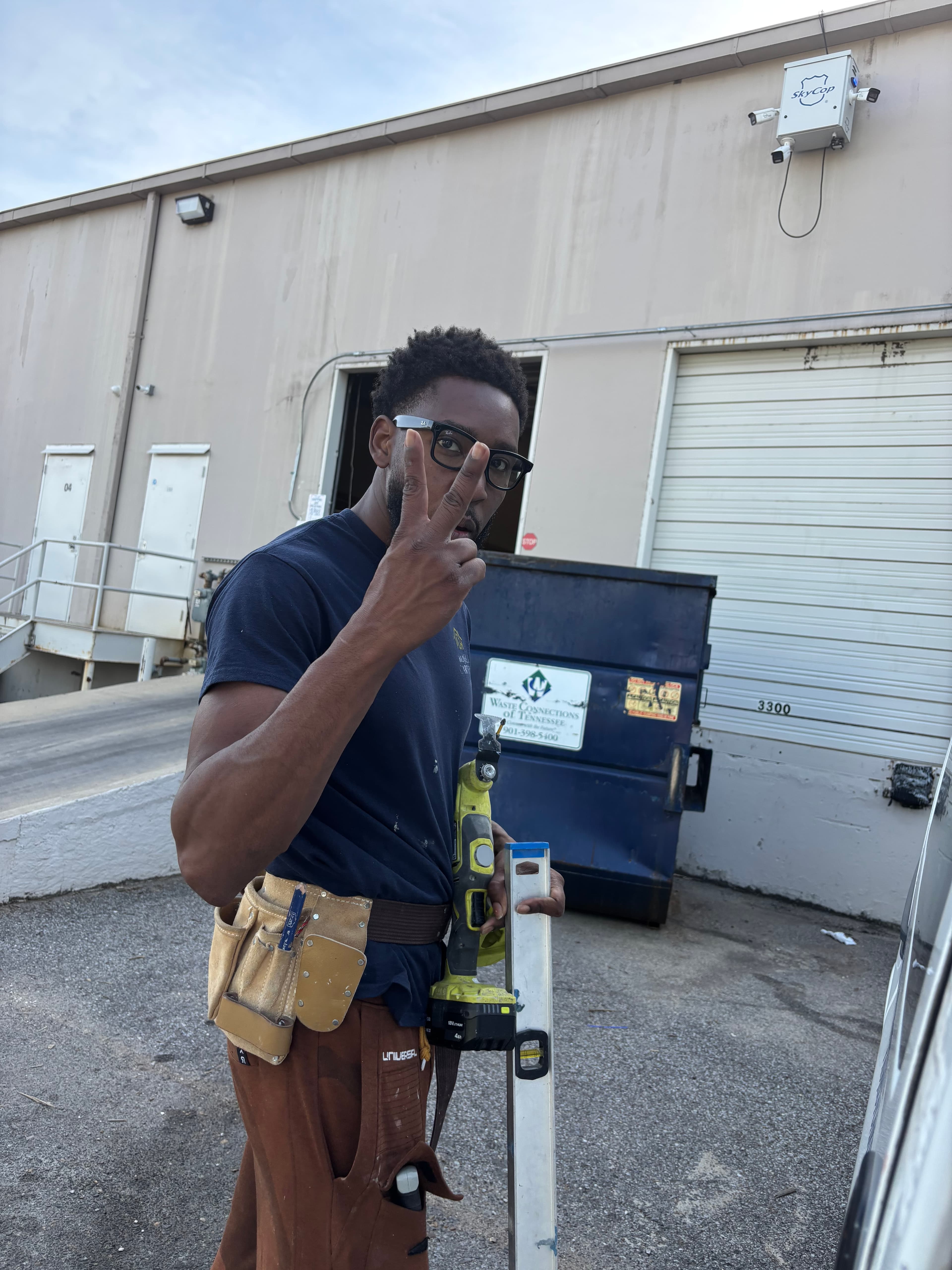 Black man with glasses and tool belt makes a peace sign holding construction tools.